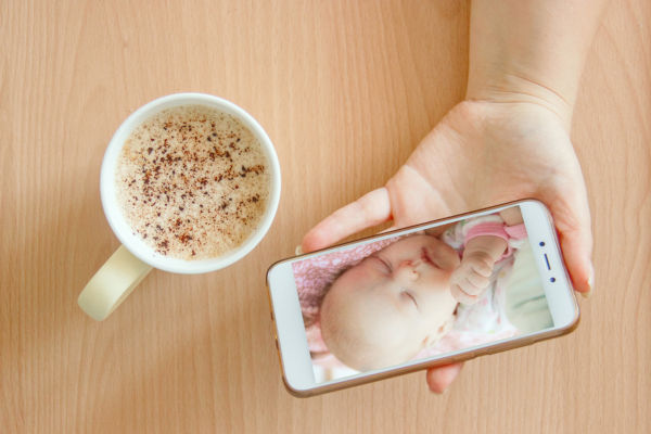 view handheld color video baby monitor. Female hands are holding a smartphone with a baby monitor app. Near hot drink.