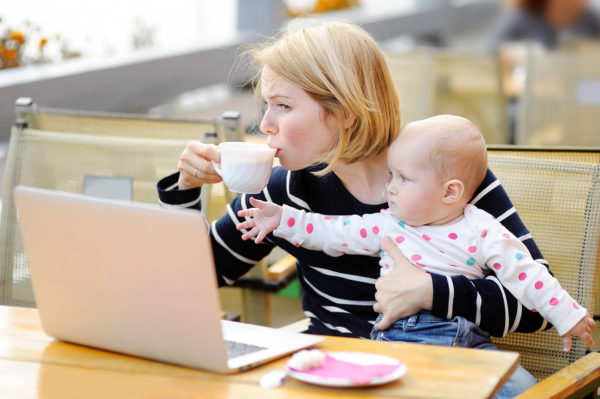 Tired young mother working oh her laptop holding daughter and drinking coffee