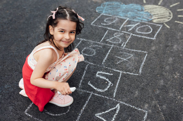 Smiling little girl drawing with chalk hopscotch on playground. Child playing the game outside. Kid wearing dress during playing hopscotch drawn on pavement. Activities for children.