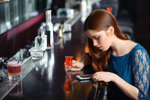 Young attractive woman looking at the screen of her phone sitting in the bar with a cocktail