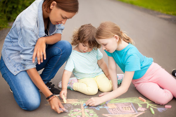 Little girls chalking at the asphalt with elder sister or nanny. Sisters friendship. Best friends children happy family. Drawing with color chalk. Happy childhood. Kids in kindergarten. Preschooler leisure time. Kids spending time without parents. Childre