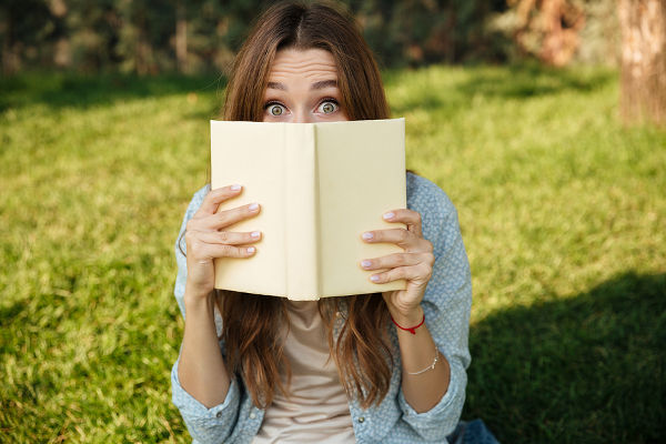 Young brunette woman sitting in park and hiding behind a book