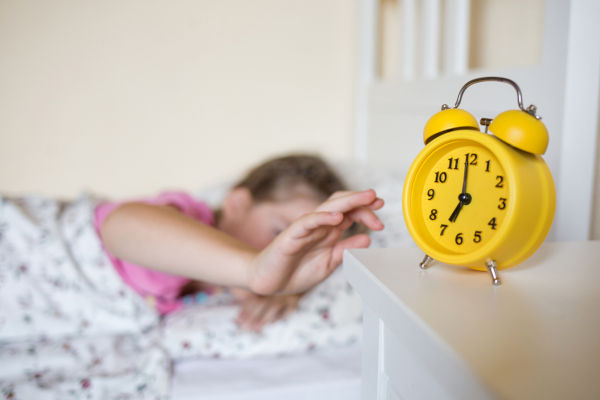 school child sleeps in bed. yellow alarm clock on nightstand in background of bed and sleeping kid in interior of room.