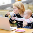 Tired young mother working oh her laptop holding daughter and drinking coffee