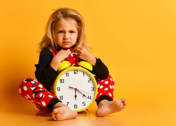 Little blond girl in red dotted pajamas sitting on floor with big alarm clock and feeling tired over yellow background. Different times of day and children schedule concept