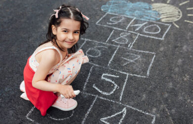 Smiling little girl drawing with chalk hopscotch on playground. Child playing the game outside. Kid wearing dress during playing hopscotch drawn on pavement. Activities for children.