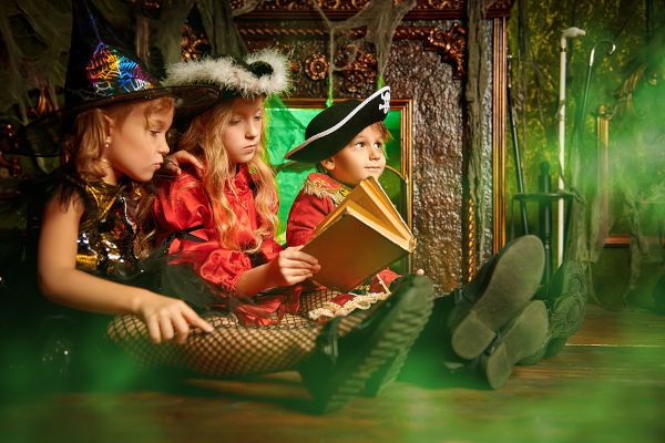 Children in carnival costumes sit together by the fireplace in an old house and read Halloween tales.