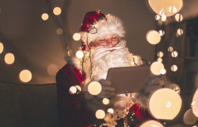 Santa Claus sitting on the couch in warm winter holidays home atmosphere, having a video call using tablet computer with Christmas lights and bokeh all around