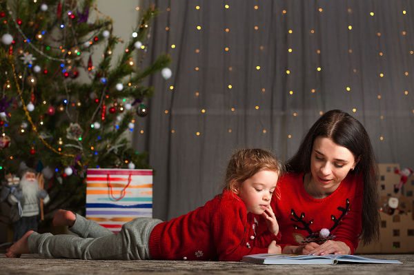 Mom and daughter in a red sweater for Christmas at the Christmas tree. The girl and her mother read books, fairy tales at the Christmas tree at home for the New Year. Stay home for the holidays.