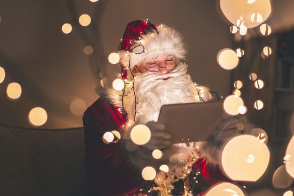 Santa Claus sitting on the couch in warm winter holidays home atmosphere, having a video call using tablet computer with Christmas lights and bokeh all around